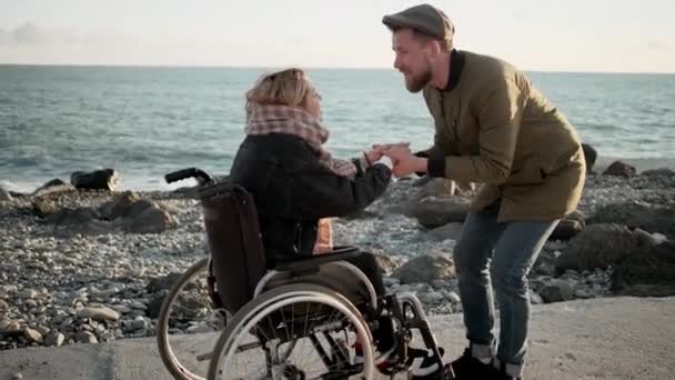 Чоловік бадьорить свою кохану жінку, сидячи в інвалідному візку на морському пляжі — стокове відео