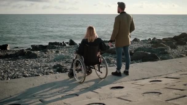 Vrouw zit in een rolstoel aan zee en houdt de hand van haar man vast. — Stockvideo