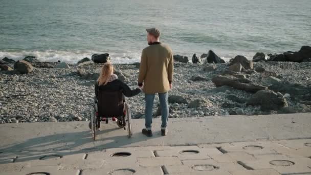 Wheelchair woman user and healthy man are holding hands and watching seascape — Stock Video