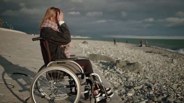 Young blonde woman with paralysis of legs is sitting in wheelchair, viewing sea — 图库视频影像