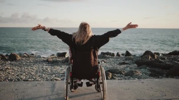 Alleen gehandicapte vrouw zit in een rolstoel en houdt hands up aan zee kust — Stockvideo