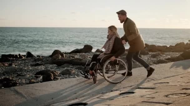 愛する男は、海の海岸で若い女性と車椅子を旋回し、喜んでいます — ストック動画