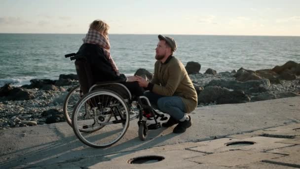 El hombre está cogido de la mano de una joven enferma en silla de ruedas y hablando con ella — Vídeo de stock