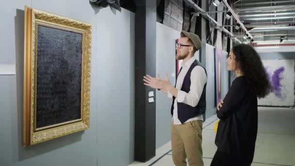 El hombre está expresando su opinión sobre la imagen en la galería de arte, la mujer está escuchando — Vídeo de stock