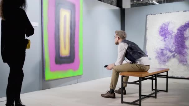 Молодой человек слушает музыку через наушники, просматривая картинку в галерее — стоковое видео
