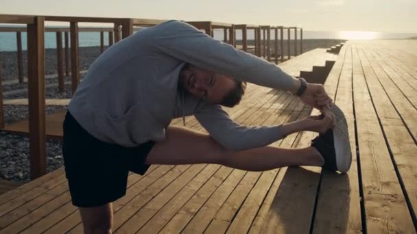 Joven hombre deportivo está haciendo ejercicio fuera en el tiempo de puesta del sol, estirando la pierna — Vídeos de Stock