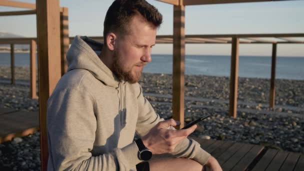 Красивый парень читает сообщения в своем смартфоне на берегу моря на закате — стоковое видео