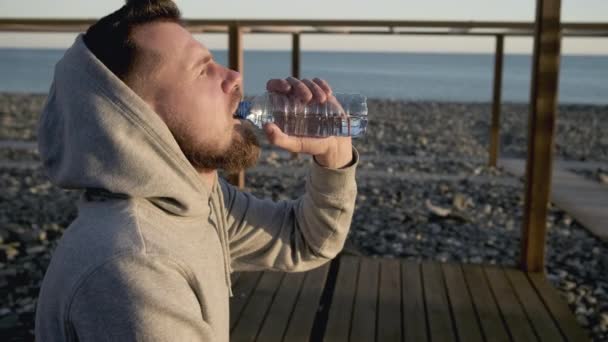 Дорослий чоловік розслабляється на морському березі, п'є воду і дихає свіжим повітрям — стокове відео