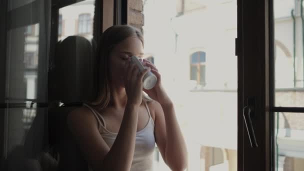 Женщина пьет чай сидя на подоконнике открытого окна — стоковое видео