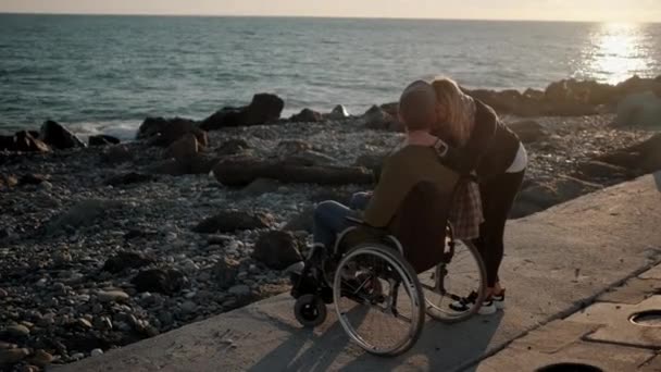 Zdravá holka si objímá nemocného přítele v kolečkovém křesle na mořském molu — Stock video