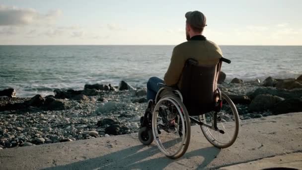 El hombre solitario está sentado en un carruaje inválido en la orilla del mar, mirando a lo lejos — Vídeos de Stock