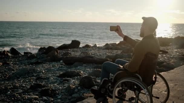 Mann sitzt in ungültiger Kutsche und macht per Smartphone Fotos und Videos vom Meer — Stockvideo