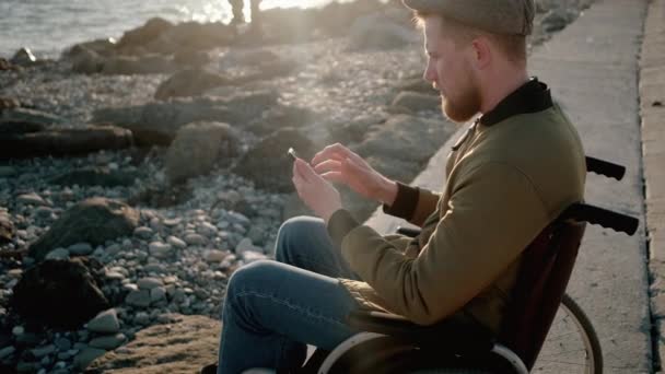 Мужчина, сидящий в инвалидной коляске на набережной, пользуется смартфоном с интернетом, стягивает — стоковое видео