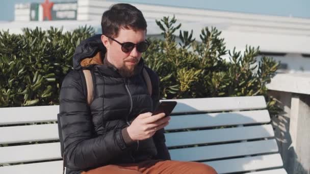 Turista en gafas de sol tipos en el teléfono inteligente sentado en el banco — Vídeo de stock