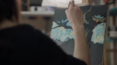 Kadın tuvale bir resim çiziyor, yağlı boyalarla, omuzdan manzarayla.