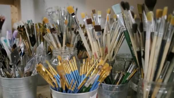 Muchas copas con pinceles artísticos están de pie sobre la mesa en el estudio de arte, panorámica — Vídeos de Stock