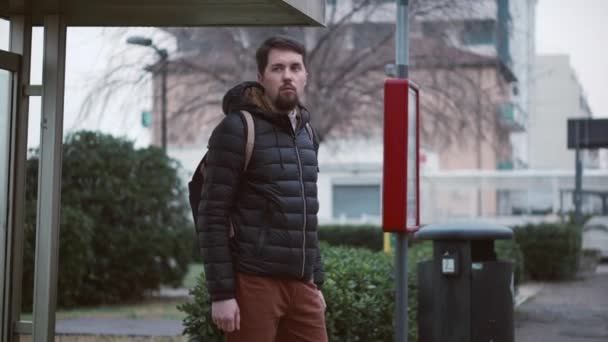 Vousatý chlap v bundě stojí na veřejné autobusové zastávce — Stock video