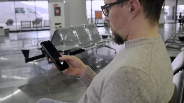 Ragazzo naviga internet in smartphone seduto in aeroporto — Video Stock