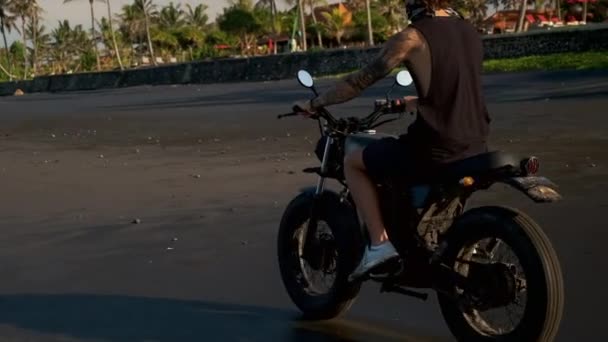 Tattooed man drives motorbike passing palm trees and huts — Stock Video