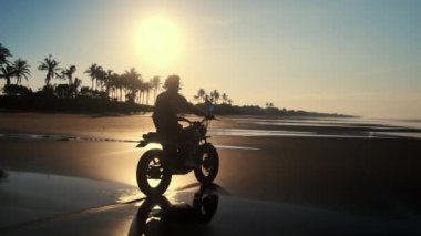 Motosikletli gün batımında deniz kıyısında at sürüyor.