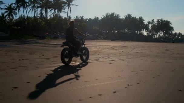 Motociclista conduciendo por la playa de arena más allá de bungalows y árboles tropicales — Vídeos de Stock