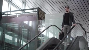 Ciddi bir adam büyük havaalanında yürüyen merdivenden iniyor.