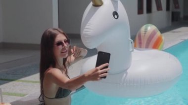 Neşeli bikinili kız havuzda tek boynuzlu atıyla selfie çekiyor.