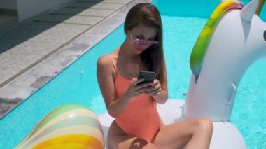 Kadın akıllı telefonuyla mesajlaşıyor havuzda şişme tek boynuzlu atın üzerinde oturuyor.