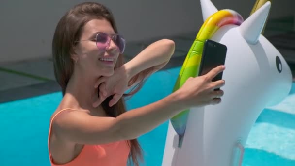 Kadın akıllı telefonuyla selfie çekiyor, şişme tek boynuzlu atta yüzüyor. — Stok video
