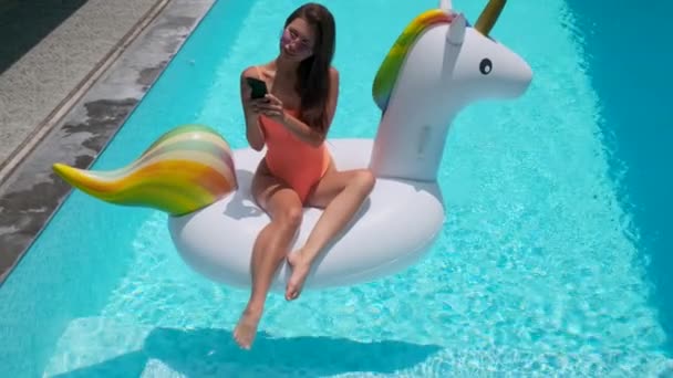 Adorável jovem relaxante na piscina com um telefone celular em um unicórnio inflável — Vídeo de Stock