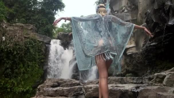 Молода жінка в літаючому одязі піднімається до водоспаду і починає свій танець — стокове відео