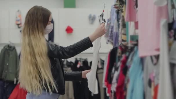 Mujer joven con máscara médica comprando camiseta en el hipermercado — Vídeo de stock