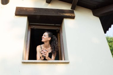 Gülümseyen kadın elinde bir fincan kahveyle bir kır evinin penceresinden dışarı bakıyor.