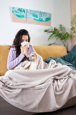 Grip ya da alerji belirtileri gösteren bir kadın evinde köpeğiyle kanepede otururken burnunu siliyor.