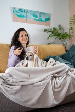 Gülümseyen bir kadın, kanepede köpeğiyle eğleniyor, televizyon izliyor ve çay içiyor.