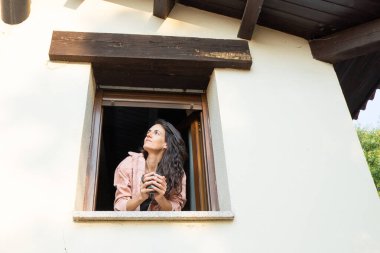 Elinde bir kupa tutan ve umutla bakan bir kadın, bir evin penceresinde sabah kahvesinin tadını çıkarıyor.