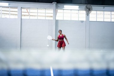 Pickleball oynayan genç bir kadın, kapalı alanda mavi bir kortta küreğiyle topa vurmak üzere.