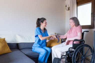 Gülümseyen hemşire tekerlekli sandalyede oturan yaşlı bir kadının kan basıncını ölçüyor ve eve sağlık hizmeti veriyor.