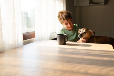 Genç bir çocuk, mutfak masasında köpeğiyle huzurlu bir sabah geçiriyor. Pencereden güneş ışığı akıyor.