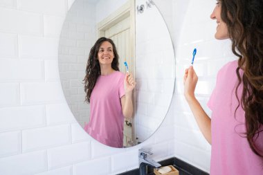 Genç bir kadın dişlerini fırçalıyor ve banyo aynasında yansımasına gülümsüyor. Günlük ağız temizliğini teşvik ediyor.