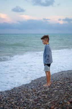 Düşünceli çocuk çakıl taşı plajında duruyor, alacakaranlıkta okyanus dalgalarını düşünüyor.