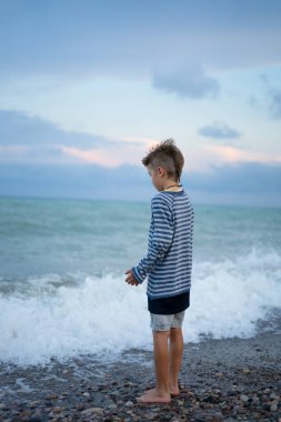 Düşünceli çocuk alacakaranlıkta çakıllı bir plajda çıplak ayakla duruyor ve önünde okyanus dalgalarının düşüşünü izliyor.