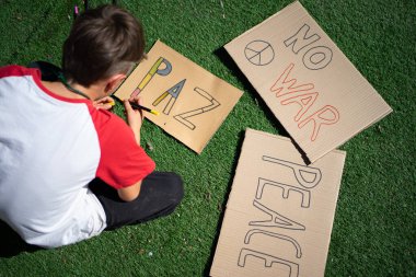 Protesto tabelaları hazırlayan genç bir çocuk. Kartonda savaş yok, çimenlerde oturuyor.