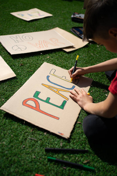 Child preparing a protest sign promoting peace and condemning war, drawing colorful letters on cardboard