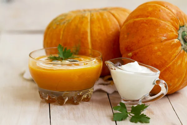 home made pumpkin cream soup with pumpkins and parsley leaves on white wooden table. pumpkin soup with sour cream and pumpkin seeds in a glass bowl. cream soup of pumpkin