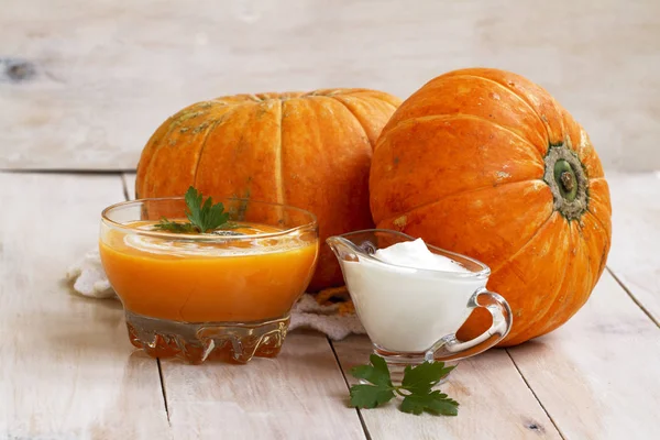 home made pumpkin cream soup with pumpkins and parsley leaves on white wooden table. pumpkin soup with sour cream and pumpkin seeds in a glass bowl. cream soup of pumpkin