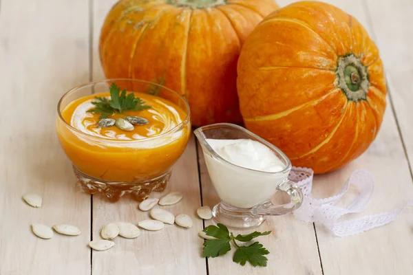 home made pumpkin cream soup with pumpkins and parsley leaves on white wooden table. pumpkin soup with sour cream and pumpkin seeds in a glass bowl. cream soup of pumpkin