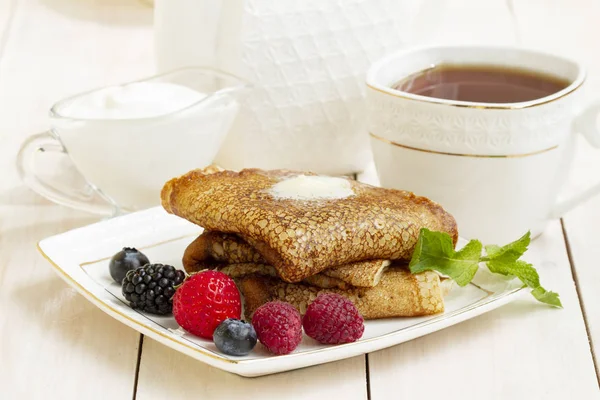 Panqueques con bayas, yogur y té en una taza de porcelana blanca. Desayuno saludable de verano, panqueques rusos clásicos caseros con baya fresca y crema agria, fondo de madera blanco claro por la mañana . — Foto de Stock