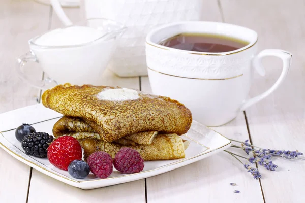 Panqueques con bayas, mantequilla y té de lavanda en taza de porcelana. Desayuno saludable de verano, panqueques rusos clásicos caseros con baya fresca y crema agria, fondo de madera blanco claro por la mañana . — Foto de Stock