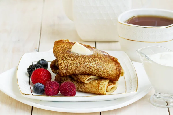 Panqueques con bayas, mantequilla y té en una taza de porcelana blanca. Desayuno saludable de verano, panqueques rusos clásicos caseros con baya fresca y crema agria, fondo de madera blanco claro por la mañana . — Foto de Stock